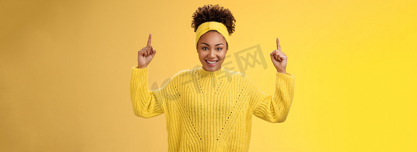 黄色促销摄影照片_兴奋可爱的女性非洲裔美国年轻 20 多岁女孩非洲发型，头戴毛衣，手指向上，广泛微笑，呈现空白促销，笑得很开心，推荐产品