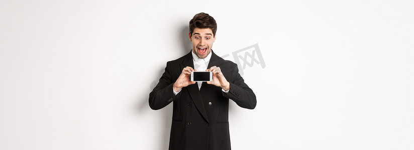 兴奋的英俊男人的肖像展示智能手机屏幕，惊讶地看到广告展示，穿着黑色西装站在白色背景上