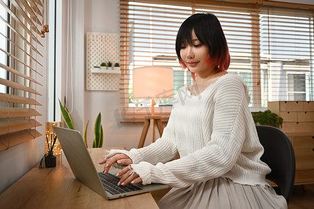 浏览互联网摄影照片_年轻漂亮的亚洲女性坐在舒适的客厅里，使用笔记本电脑浏览互联网并在社交网络上查看视频
