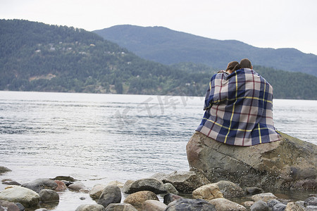 坐在海边摄影照片_一对裹着毯子坐在海边岩石上的夫妇
