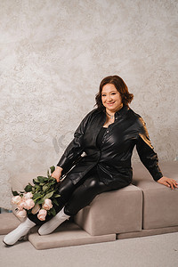 一位身穿黑色皮衣的时尚成年女性坐在室内，里面放着一束粉红玫瑰