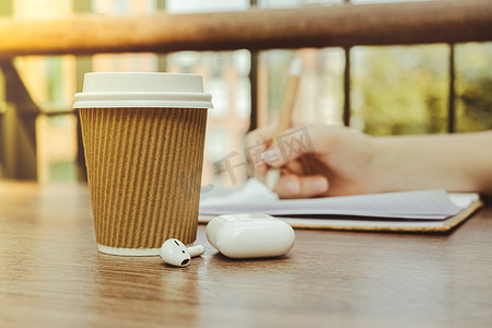 工艺回收纸杯中的咖啡无法辨认的女人用无线耳机在纸质笔记本上写字。