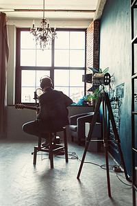 男歌手坐在凳子上，戴着耳机，吉他在家庭录音室录制曲目