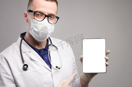 年轻英俊的聪明医生，戴着灰色背景的外科卫生防护口罩，手里拿着一台带有空白样机屏幕的平板电脑。