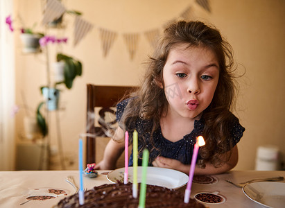 儿童节庆祝摄影照片_可爱的女婴，长着美丽的长卷发，穿着优雅的海军服，吹灭生日蛋糕上的蜡烛