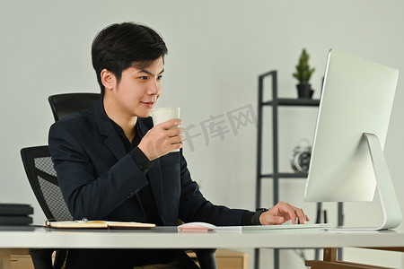 身穿西装、微笑的亚洲男子专属经理喝着热咖啡，在电脑显示器上检查商业电子邮件