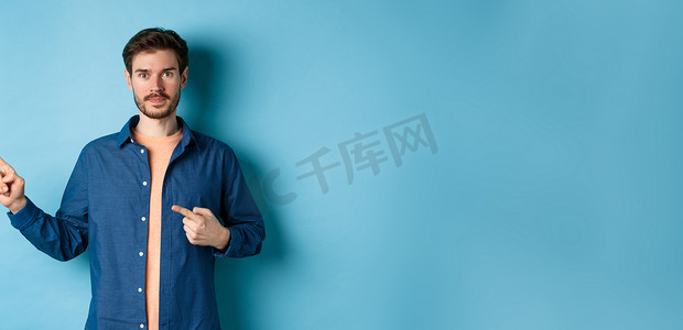 穿着衬衫的英俊年轻人，用手指指着促销片，展示标志，站在蓝色背景上