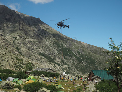 法国，科西嘉岛，科西嘉阿尔卑斯山，2017年6月19日：直升机在著名的远足小径GR20、岩石、帐篷、树木和蓝天背景上为山地营地Refuge de Pietra Piana运送物资