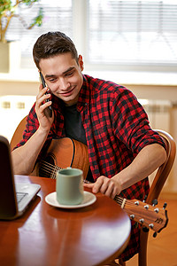 放松的年轻人在家弹吉他，用笔记本电脑，喝茶，笑着打电话
