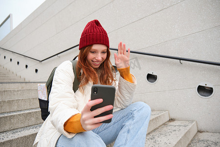 快乐的女大学生通过视频聊天智能手机应用程序与朋友交谈，坐在户外楼梯上使用她的手机应用程序，对着电话摄像头挥手