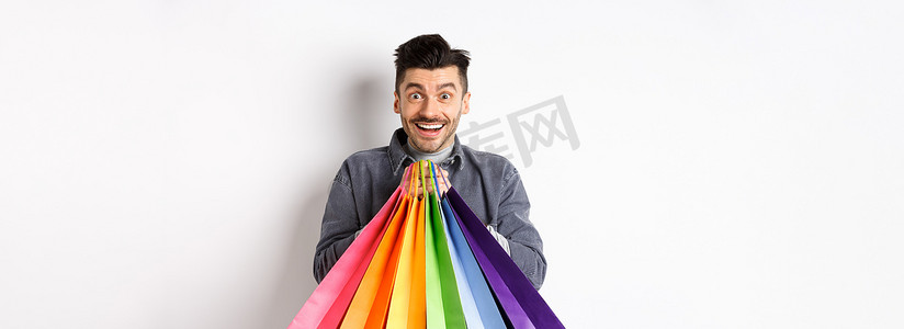 站在白色背景下，兴奋的微笑男子拿着彩色购物袋，对商店里的折扣感到高兴