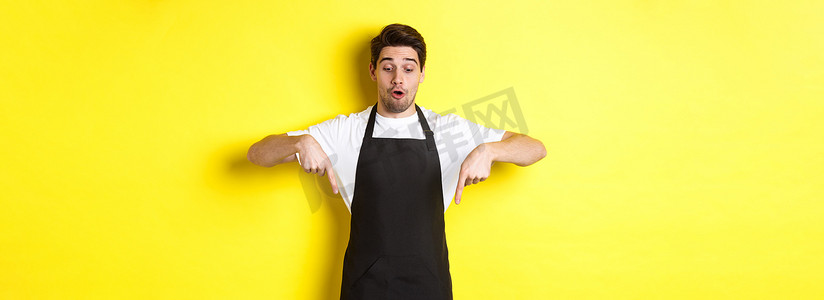 身穿黑色围裙的男性咖啡师惊讶地查看促销优惠，手指向下指着徽标横幅，站在黄色背景上