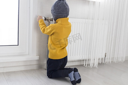 毛线帽子摄影照片_一个穿着黄色毛衣、戴着灰色帽子的小孩跪在带恒温器的加热器附近。