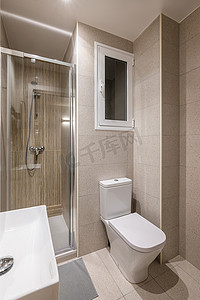 多功能小浴室配有时尚的米色瓷砖、玻璃淋浴门、洗脸盆、卫生间和窗户。
