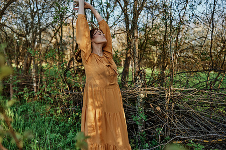 绿色树苗条摄影照片_一位美丽、苗条的长发女子走在树旁的树荫下，穿着橙色长裙，享受天气和周末，高兴地举起双手