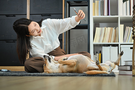 狗狗手机摄影照片_美丽的女人使用智能手机与可爱的狗躺在家里的地板上自拍。