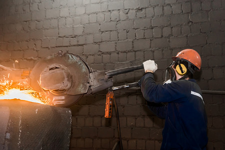 一名戴着防护头盔、呼吸器、工作服的男工人在工厂车间管理带有飞火花的铸铁混凝土管的重型研磨设备