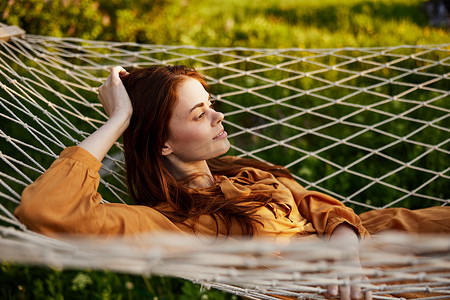 一个快乐的女人躺在网状吊床上，头枕在手上，幸福地微笑着看着别处，在夕阳的光芒下享受温暖的一天，躺在橙色的裙子上