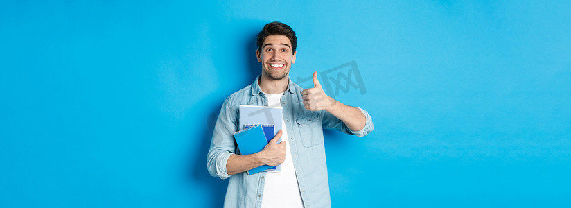 快乐的年轻男模特拿着笔记本，高兴地竖起大拇指，微笑着推荐课程，站在蓝色背景上
