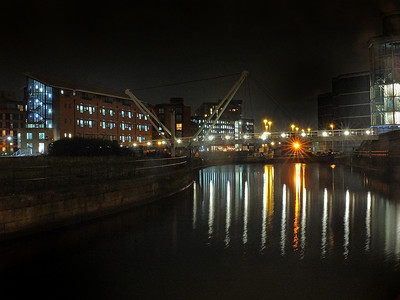 路人甲路人乙摄影照片_骑士路人行桥在夜间穿过利兹克拉伦斯码头的船闸入口处的河流，建筑物和灯光倒映在水中