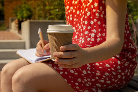 穿着红色连衣裙、用工艺纸杯喝咖啡、在木凳上写感恩日记的无法辨认的年轻女子。