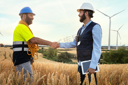 快乐的男性电气工程师和微笑的维护工人在风力涡轮机农场握手。