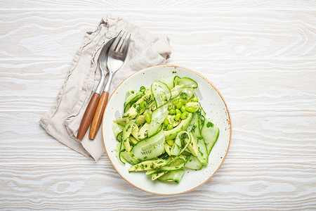 健康的纯素绿色鳄梨沙拉碗，陶瓷板顶视图上有切片黄瓜、毛豆、橄榄油和香草，白色木质质朴的桌子背景