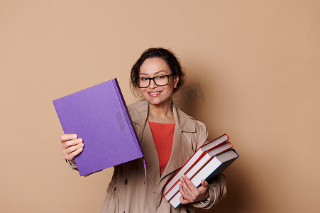 教育家摄影照片_戴着眼镜的美丽女教师教育家教授，一边拿着米色背景的书，一边对着镜头微笑
