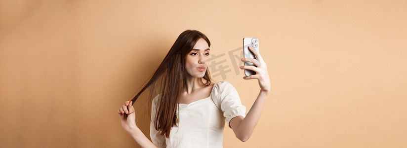 时尚的年轻女性玩头发，在智能手机上自拍，为社交媒体拍照，站在米色背景上