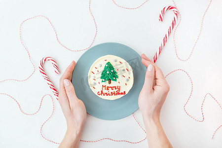 一个女孩拿着一个盘子，上面有一个圣诞蛋糕，上面刻着“圣诞快乐”，上面装饰着糖霜，白色背景上有棒棒糖和一根红线作为礼物。