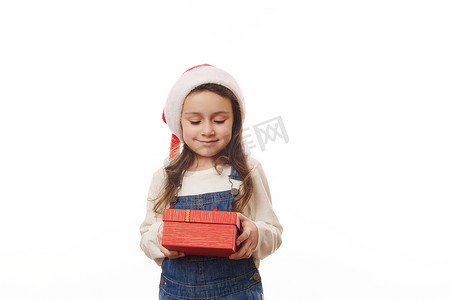 白色背景中戴着圣诞帽的可爱小女孩的肖像，手里拿着一个红色礼盒，为圣诞节带来惊喜