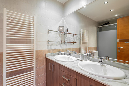 浴室的天花板灯发出明亮的光线，冷光束反射在水槽闪亮的白色表面和干净的大镜子上，镜子上显示木制闭门淋浴间加热散热器。