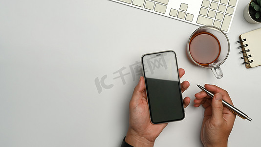 手机办公办公摄影照片_上面是男人双手拿着智能手机放在办公桌上的视图。