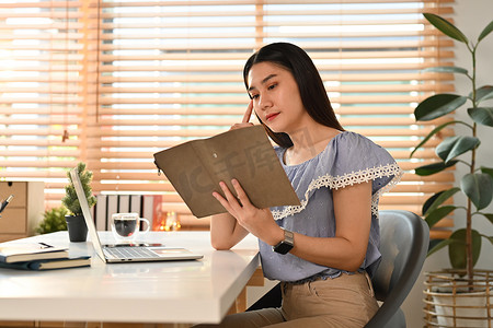 美丽的亚洲女企业家坐在舒适的工作场所，在规划师中阅读笔记