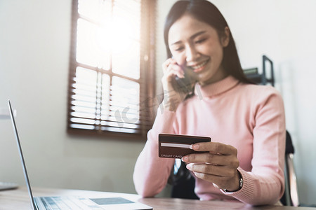 确认支付摄影照片_持有信用卡的女性通过电话联系客户服务确认购买，微笑的亚洲女孩在家里通过智能手机对话付款。