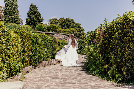 公园后景中，一位长着棕色头发和白色长裙的漂亮女人沿着美丽的灌木丛小路奔跑