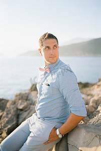 微笑的男人坐在海边的岩石海岸上，双手插在口袋里