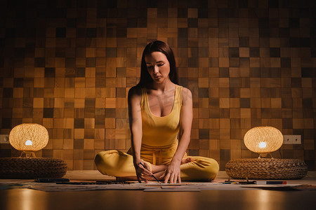 一个穿黄衣服的女孩坐在莲花位置，在家里晚上的灯光下在纸上画画