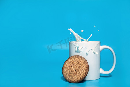 燕麦饼干落在一个白色杯子里，里面有牛奶，蓝色背景上溅着水花，有复制空间、正常健康的谷物饼干、乳制品