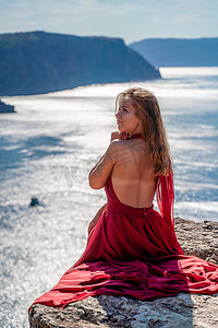 一个穿着红色长裙、头发飘逸的女孩坐在海上的一块岩石上。