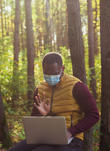戴着医用口罩的非洲裔美国人坐在城市公园的长凳上，在户外与朋友在线进行笔记本电脑视频通话。