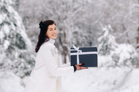 冬季服装女摄影照片_在冬季森林里，一位身穿白色西装的时尚女​​性手里拿着新年礼物。