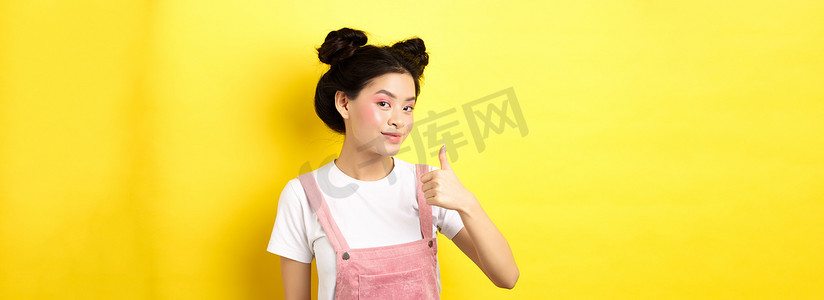 美丽的亚洲女孩，妆容亮丽，竖起大拇指，满意地微笑，推荐促销，黄色背景