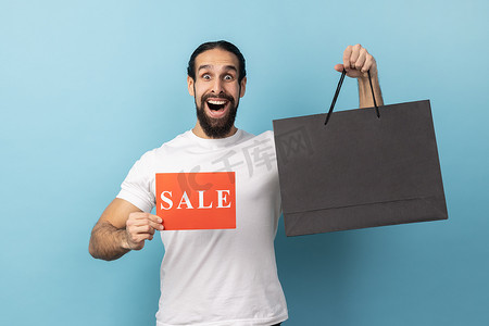 低价团购会摄影照片_震惊的男子拿着购物袋，展示销售卡，在商场里宣传低价。