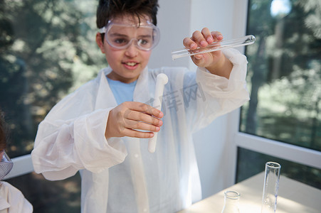 化学实验室穿着摄影照片_穿着实验室外套和护目镜的学童手中有溶液的试管，进行化学实验