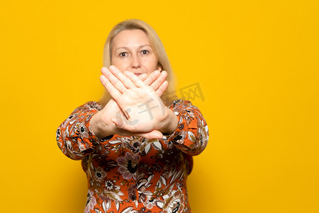 禁止手势摄影照片_40 多岁的金发美女，穿着一件带图案的毛衣，在镜头前交叉手掌做出禁止手势，孤立在黄色工作室背景中。