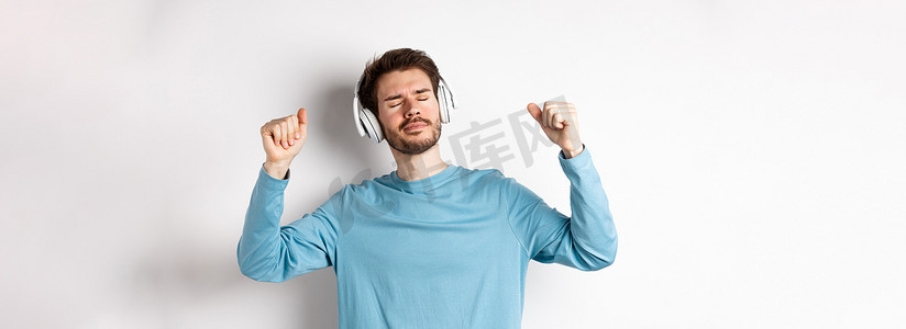 快乐的帅哥戴着耳机随着音乐跳舞，听歌微笑，站在白色背景上