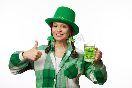 戴着三叶草耳环和派对帽的时尚女性拿着一杯爱尔兰绿啤酒，在镜头前竖起大拇指