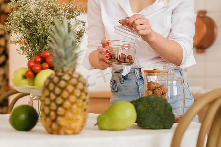女主人的手拿着罐子里的坚果，前景是厨房桌子上的蔬菜和水果的特写镜头。