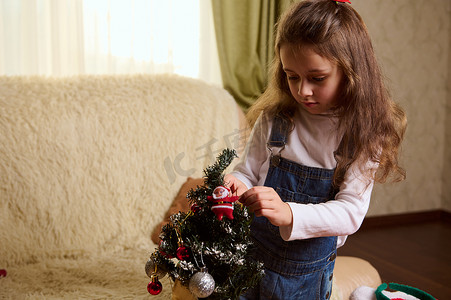 儿童拿玩具摄影照片_可爱的小女孩拿着闪亮的球，同时在舒适的家庭内部的圣诞树上挂着装饰性的圣诞玩具。